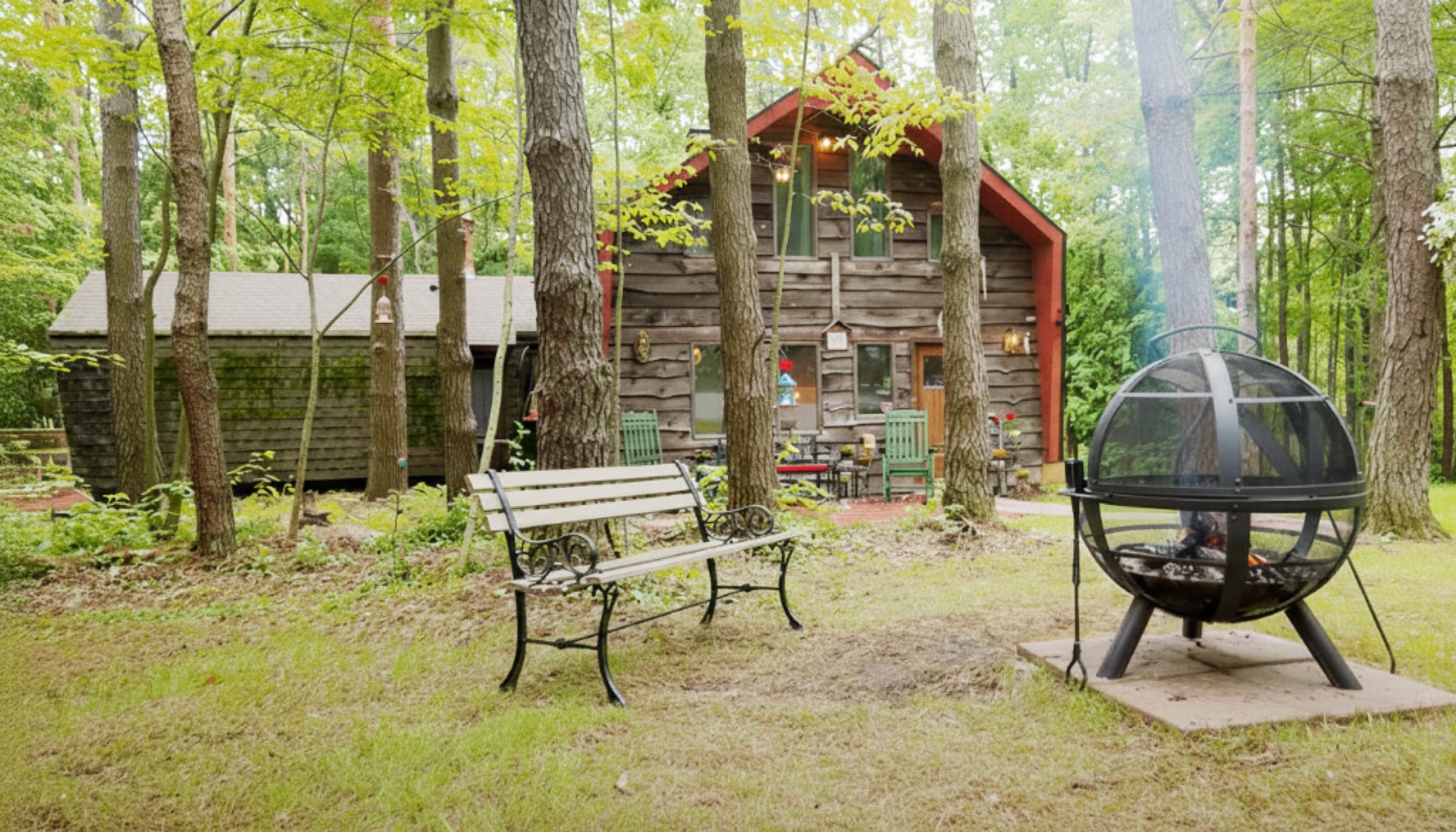 A timber cabin nestled in green forest, with a bench and fire pit out front